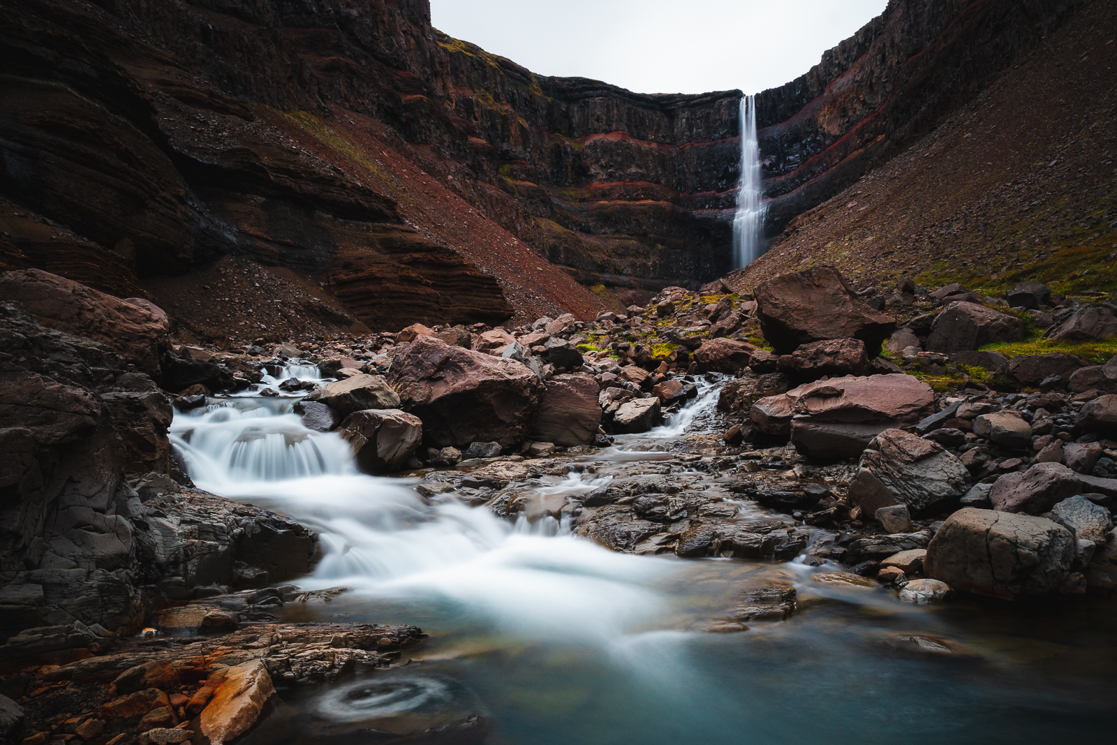 A moody little waterfall framed by epic cliffs and red rocks, doing its best impression of an emo postcard from East Iceland.