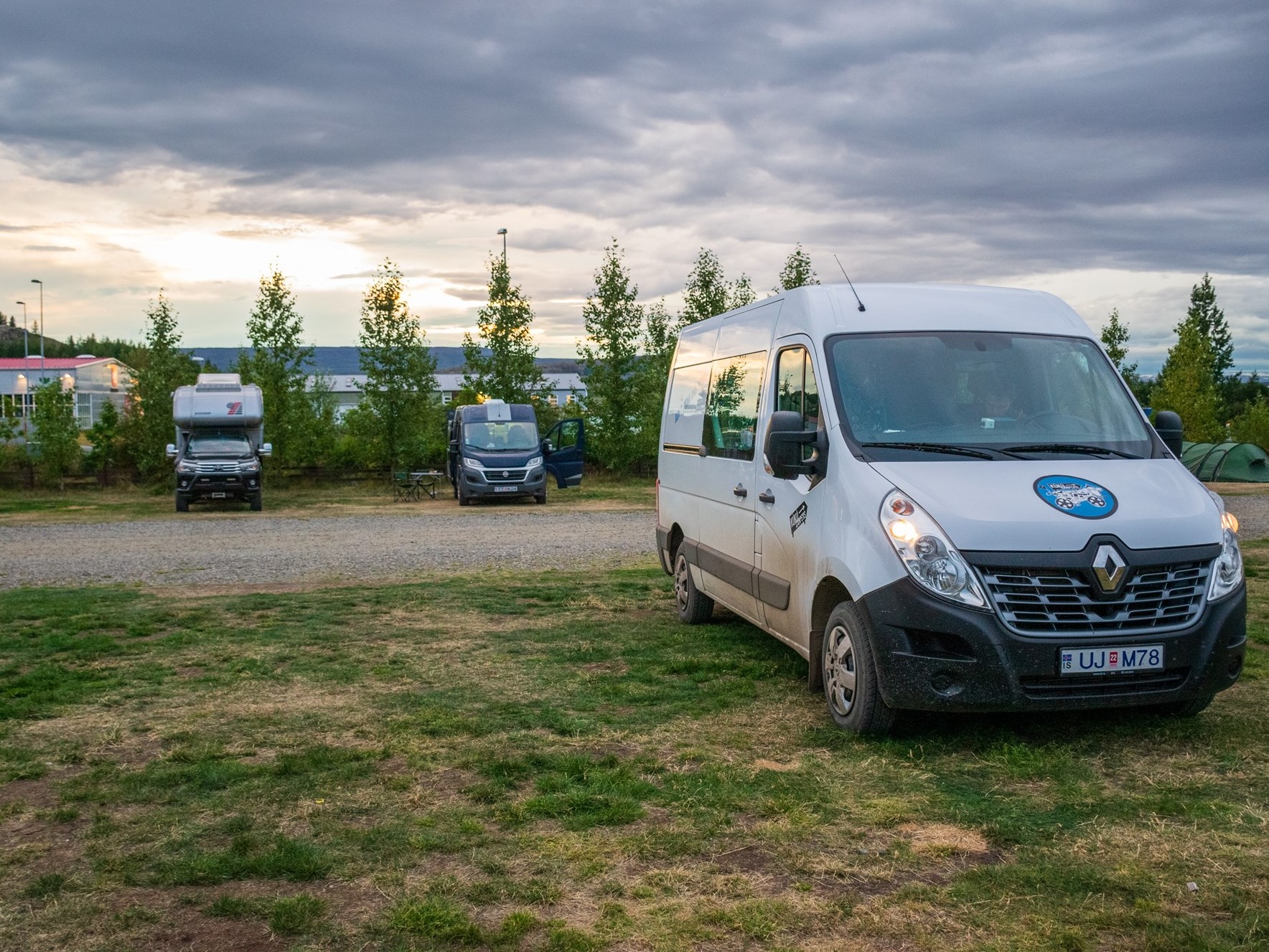 A KuKu camper van parked at Camp Egilsstaðir with a backdrop of evenly spaced trees and an open sky – a quiet and comfy home base in East Iceland.