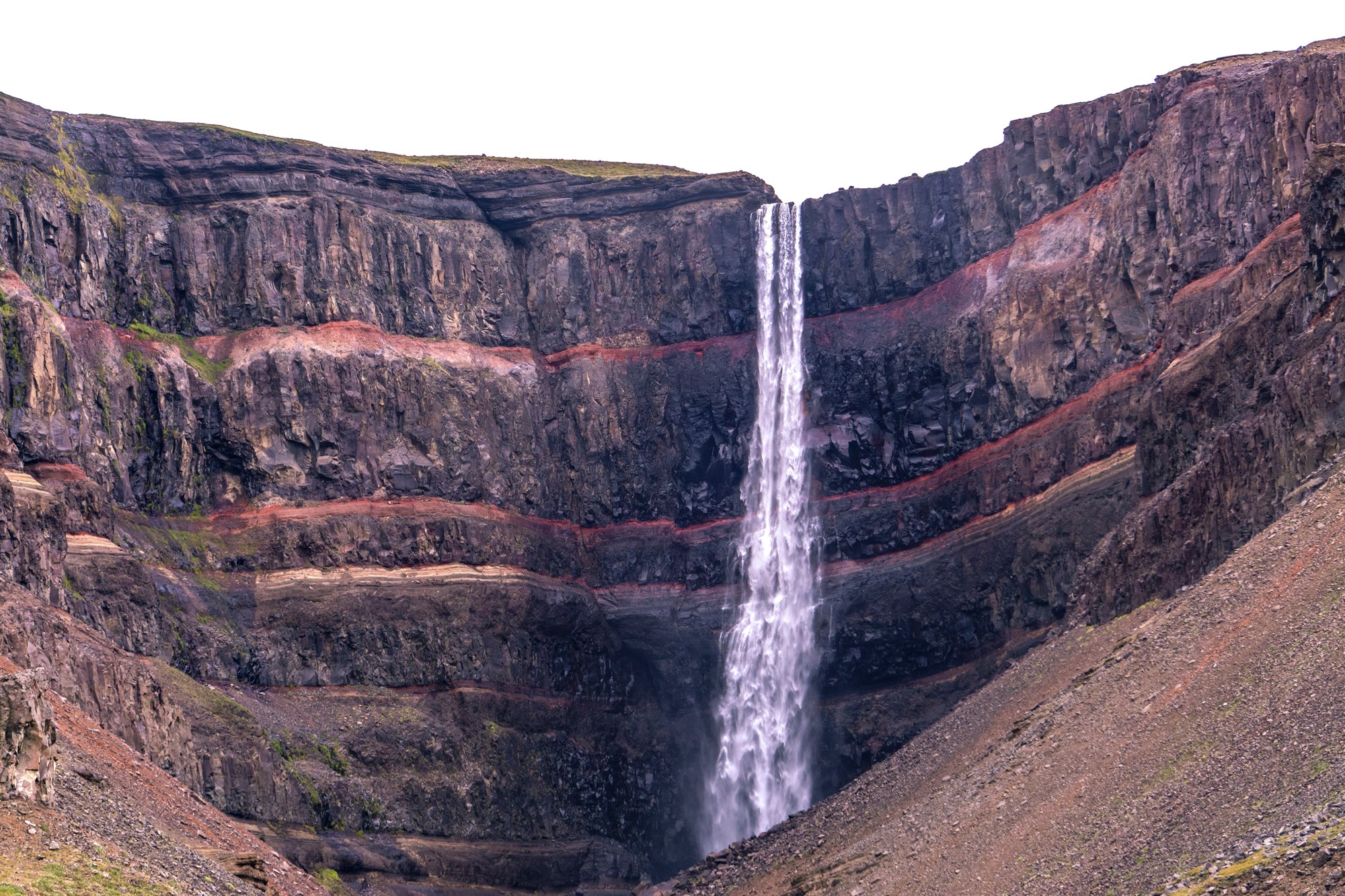 A distant view of Hengifoss waterfall cascading through vibrant red basalt layers, a striking geological wonder in East Iceland