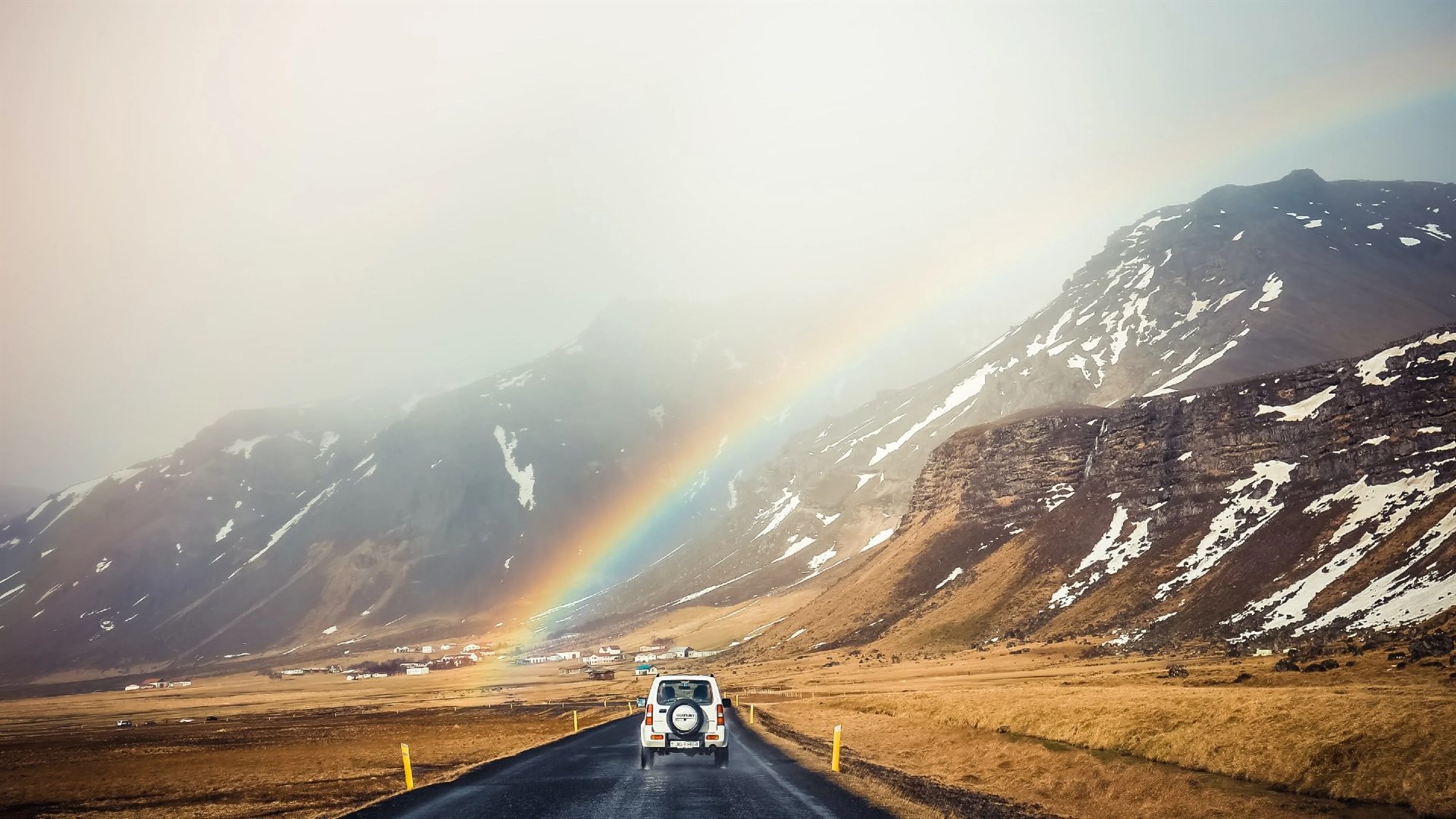 A campervan drives toward a rainbow through Iceland's stunning mountain scenery.