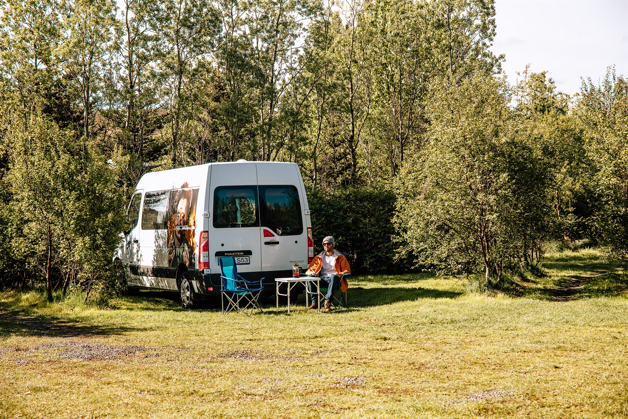 Forest Chill Spot at Mosskógar A camper van parked between tall trees with two travelers relaxing outside – peaceful vibes in one of Reykjavík’s best-located campsites.