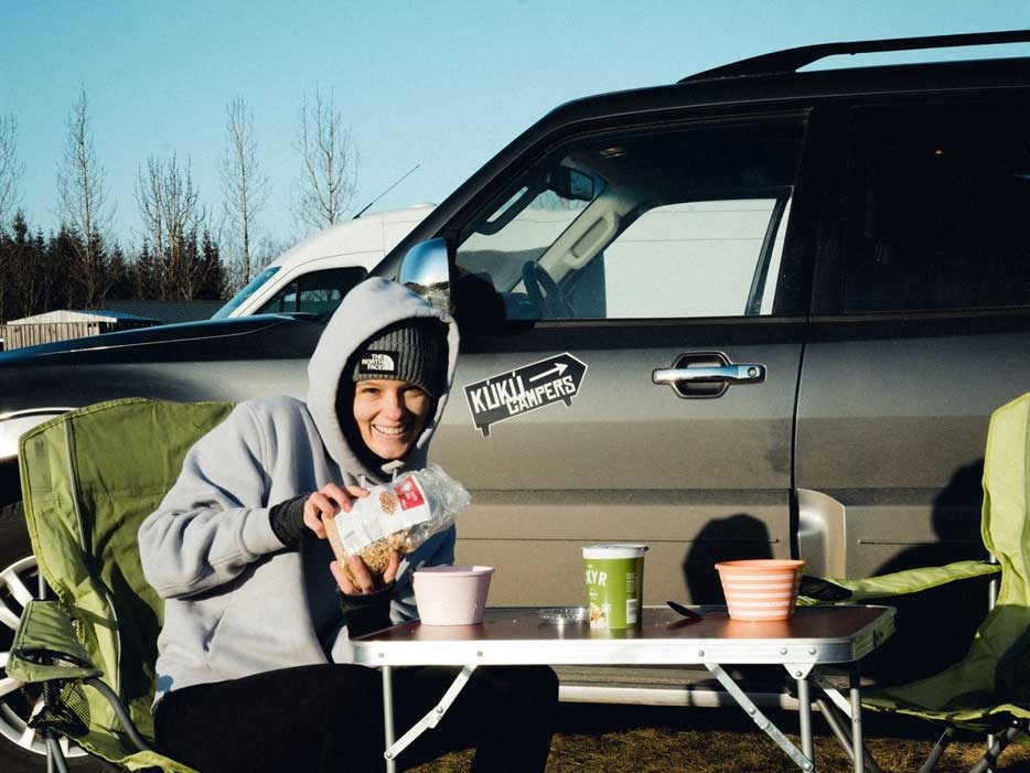 A happy traveler prepares breakfast next to a KuKu campervan, enjoying the freedom of the open road and Icelandic skies.