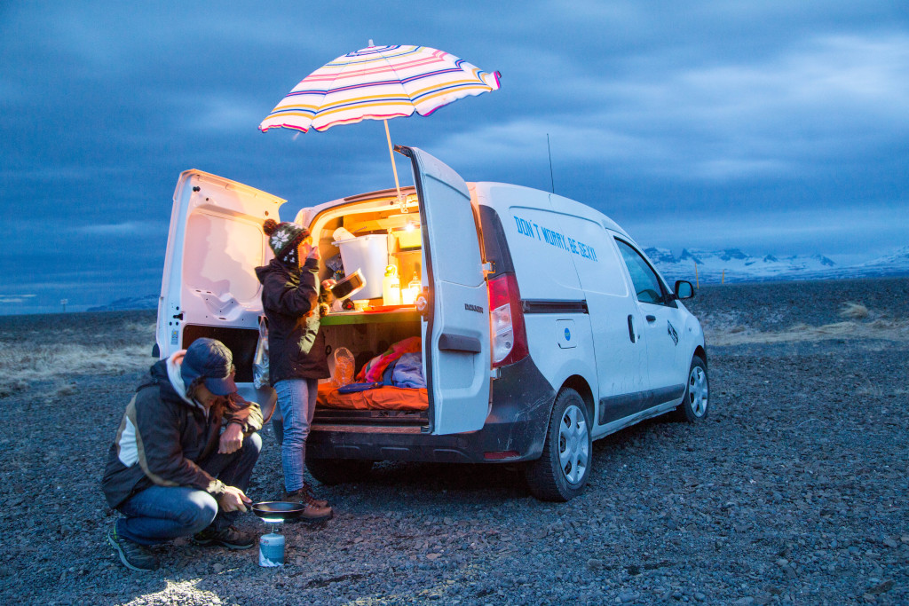 A couple cooking beside their KuKu campervan under a striped umbrella during an Iceland road trip – perfect example of van life freedom and creativity in unpredictable weather.