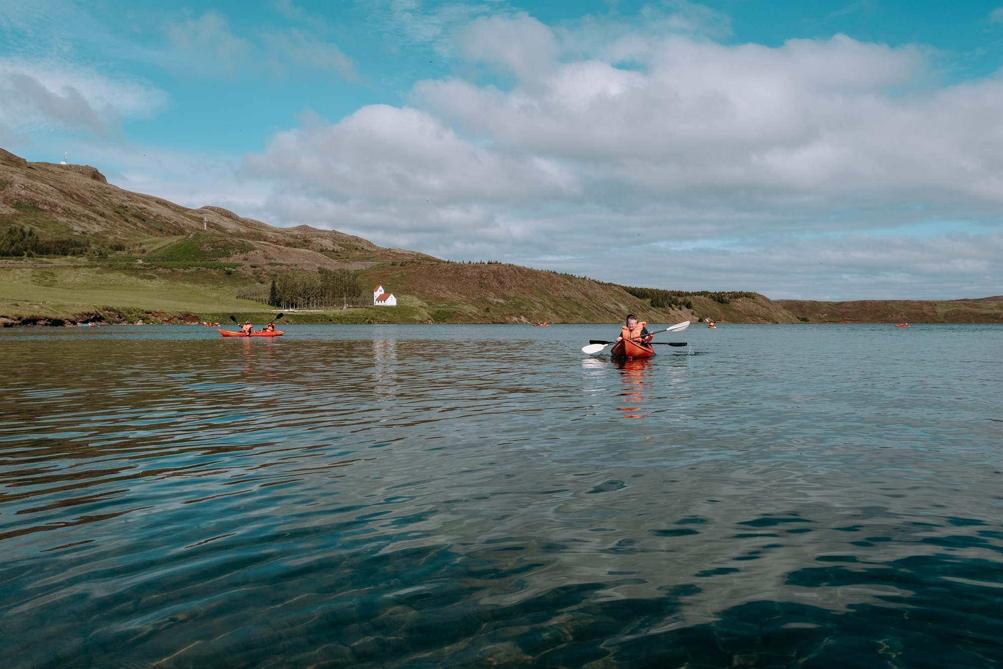 Paddling Around Úlfljótsvatn Two people kayaking on the calm waters of Lake Úlfljótsvatn, surrounded by soft hills and a red-roofed church in the distance – summer vibes at their finest in South Iceland.